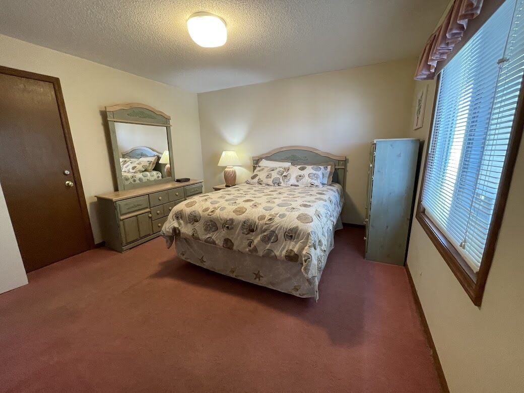 A bedroom with a bed, dresser, mirror, lamp, and window.