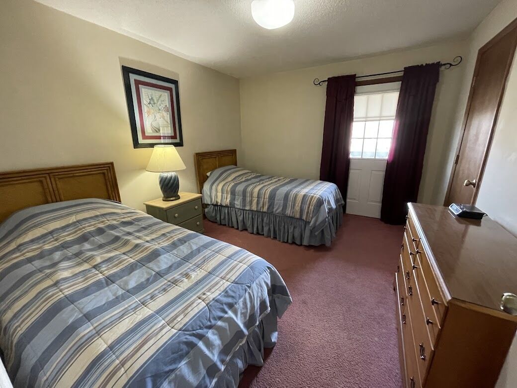 Une chambre avec deux lits, une commode, une lampe, un tableau encadré et une fenêtre avec des rideaux.