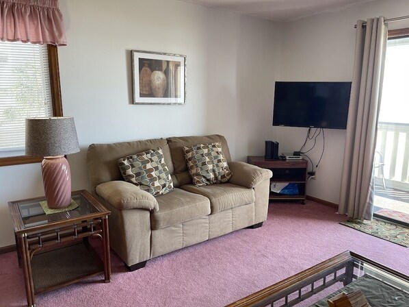 A living room with a beige sofa, patterned cushions, a glass coffee table, a TV, and a window with curtains.