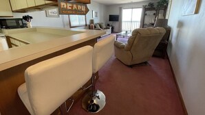A kitchen with a bar area, a beige armchair, and a television in the living room. - Surfside Landing 101 - Oceanfront in Surfside Beach (Surfside Beach)