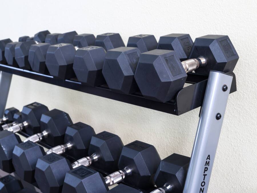 A set of black hexagonal dumbbells on a rack with a metallic frame, labeled "AMPTON."