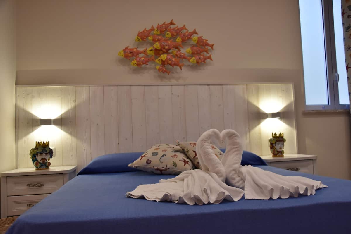 A bedroom with a blue bedspread, two nightstands with decorative items, a wall-mounted light fixture, and a floral wall decoration.