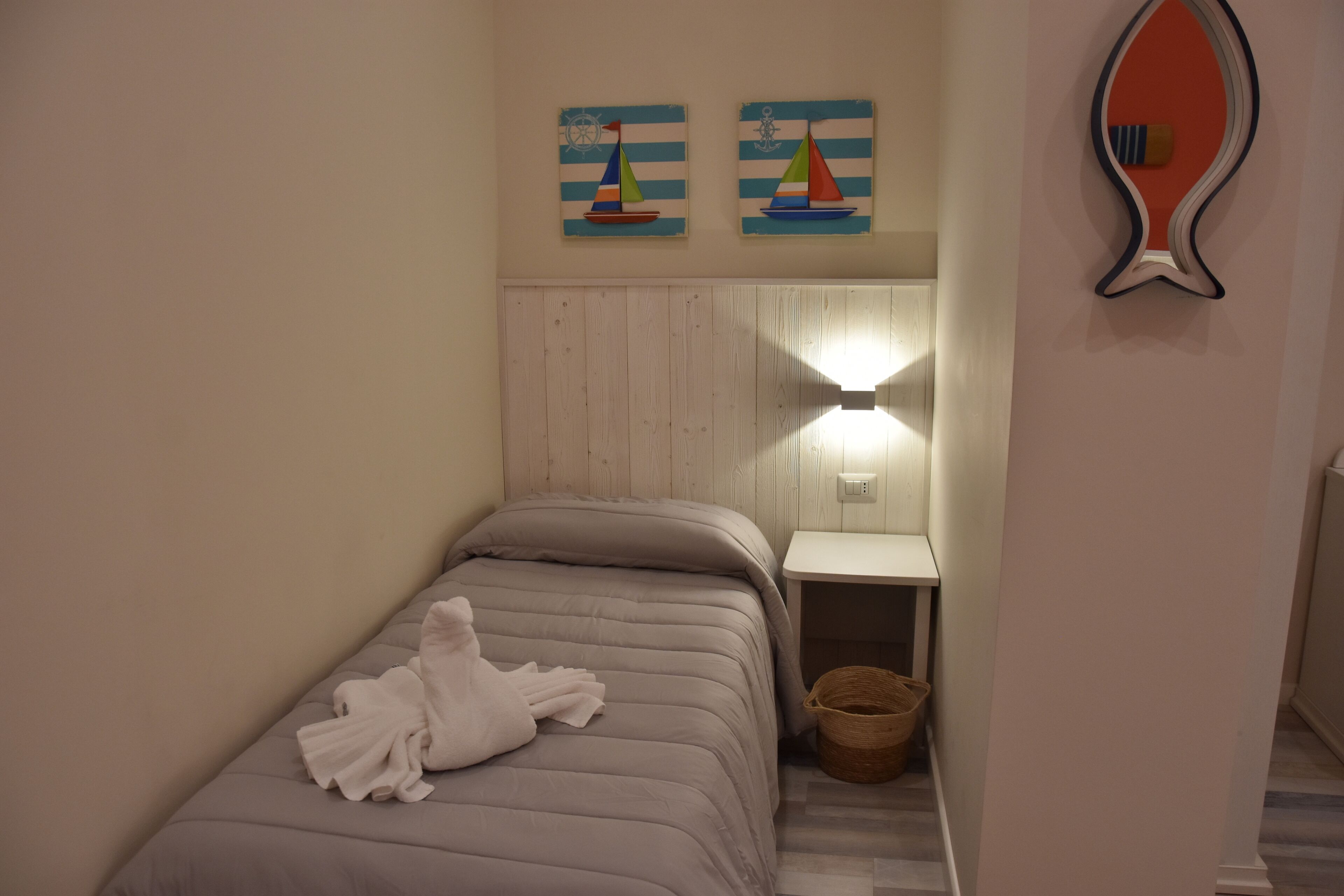 A small, neatly made bed with a white towel, a wooden headboard, and a wall-mounted nightstand with a lamp. There are two paintings of sailboats on the wall above the bed.