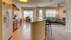 A spacious kitchen with a central island, wooden cabinets, and a built-in refrigerator. There is a dining area with a table and chairs, and a living room with a sofa and a coffee table.
