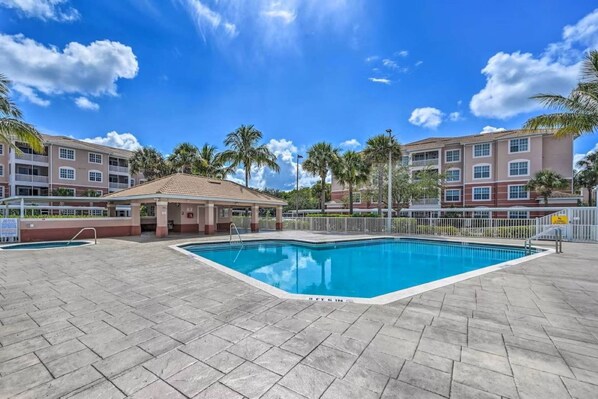 A swimming pool area with a covered seating area, surrounded by palm trees and a multi-story building with balconies. - South Fort Myers Gated Community! Heated Pool & Hot Tub! Close to Beaches, Shopping & Dining! (Fort Myers)