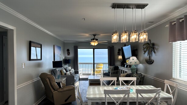 A modern living room with a dining area, large windows with a view, and a ceiling fan. - Sea Shadow 306 - Oceanfront in Surfside Beach (Surfside Beach)
