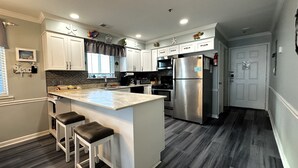 A modern kitchen with white cabinets, a stainless steel refrigerator, and a marble island with bar stools. - Sea Shadow 306 - Oceanfront in Surfside Beach (Surfside Beach)