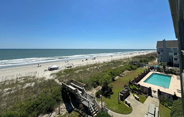 A beach view from a balcony with a swimming pool and outdoor seating.