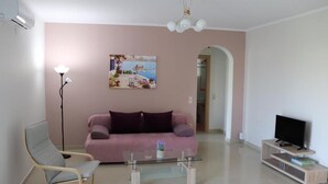 Ett vardagsrum med en rosa soffa, ett soffbord i glas, en modern stol, en tv på ett träställ och en tavla på väggen.