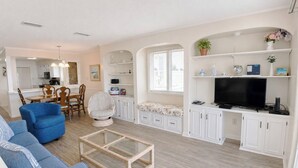 Un salon moderne avec un canapé, un fauteuil, une table basse, un téléviseur et une cuisine en arrière-plan.