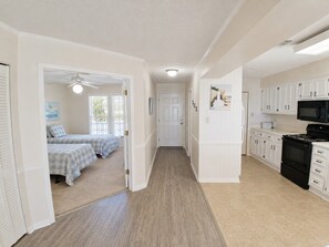 A hallway leading to a bedroom with two beds, a kitchen with white cabinets and a black stove, and a ceiling fan.