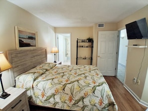 Une chambre à coucher comprenant un lit, une table de chevet, une lampe, un téléviseur fixé au mur et une porte donnant sur une autre pièce.
