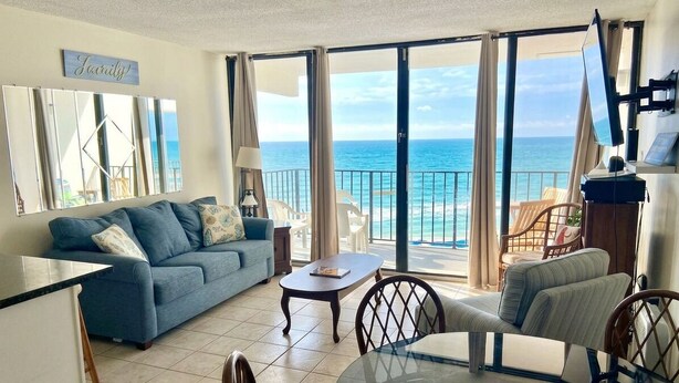 Ein Wohnzimmer mit einer Couch, einem Glastisch und Stühlen, ein Balkon mit Meerblick und einem an der Wand befestigten Flachbildfernseher.
