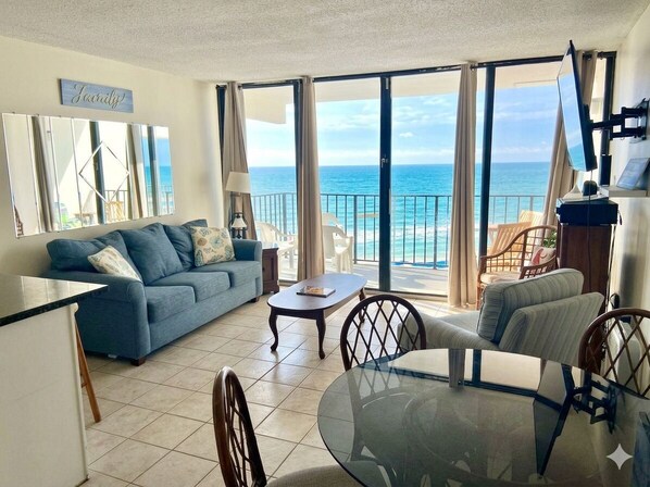 A living room with a sofa, a glass table, and chairs, a balcony with a view of the ocean, and a flat-screen TV mounted on the wall. - Royal Garden Resort 214 - Oceanfront in Garden City Beach (Garden City)