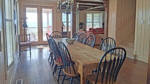 A spacious dining area with a long wooden table and black chairs, a chandelier, and a view of the ocean through large windows. - Second Creek - Oceanfront in Garden City Beach (Garden City)