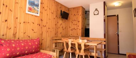 Een houten interieur met een bank, een eettafel met stoelen, een aan de muur bevestigde televisie en een deur.