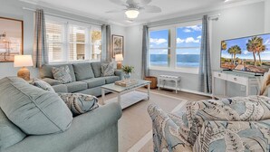 Un salon avec un canapé gris, une table basse et un téléviseur posé sur un meuble. De grandes fenêtres sont ornées de rideaux, un ventilateur de plafond est installé au plafond et un tableau encadré est accroché au mur.