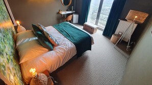 Un dormitorio con cama, lámparas de noche, escritorio, silla, mesita y espejo, además de vistas a través de un ventanal amplio.