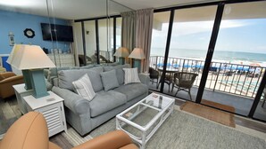 Un soggiorno con divano, tavolino e lampade. È presente un balcone con vista sulla spiaggia e sull'oceano.