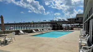 A pool area with lounge chairs and umbrellas, a pier in the background, and a building with large windows. - Sand Dollar 101 - Oceanfront in Garden City Beach (Garden City)