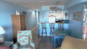 A living room with a sofa, a chair, a dining table with chairs, a kitchen area with a refrigerator and microwave, and a wooden cabinet. - Sand Dollar 101 - Oceanfront in Garden City Beach (Garden City)