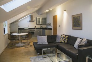 A modern apartment with a dining area, a black leather sofa, a glass coffee table, and a kitchen in the background.