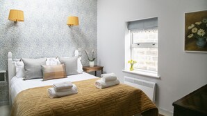 A neatly made bed with a brown quilt, two wall-mounted lamps, a window with a view of flowers, and a painting on the wall.