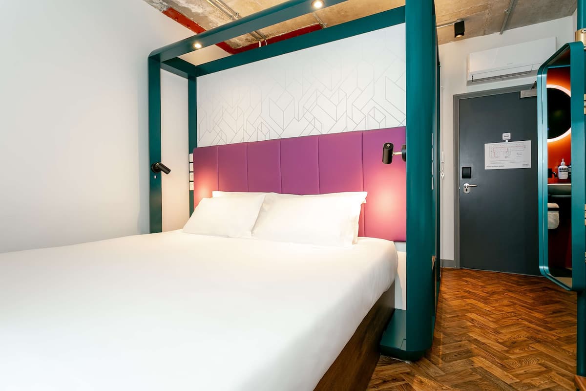 A modern hotel room with a teal-framed bed, white linens, and a purple headboard.