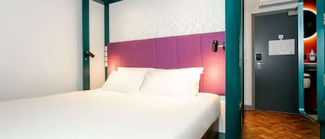 Une chambre d’hôtel moderne avec un lit doté d’un cadre de couleur turquoise, du linge de lit blanc et une tête de lit violette.