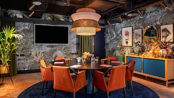 A modern dining area with a round table, orange chairs, a large pendant light, and a wall-mounted TV.