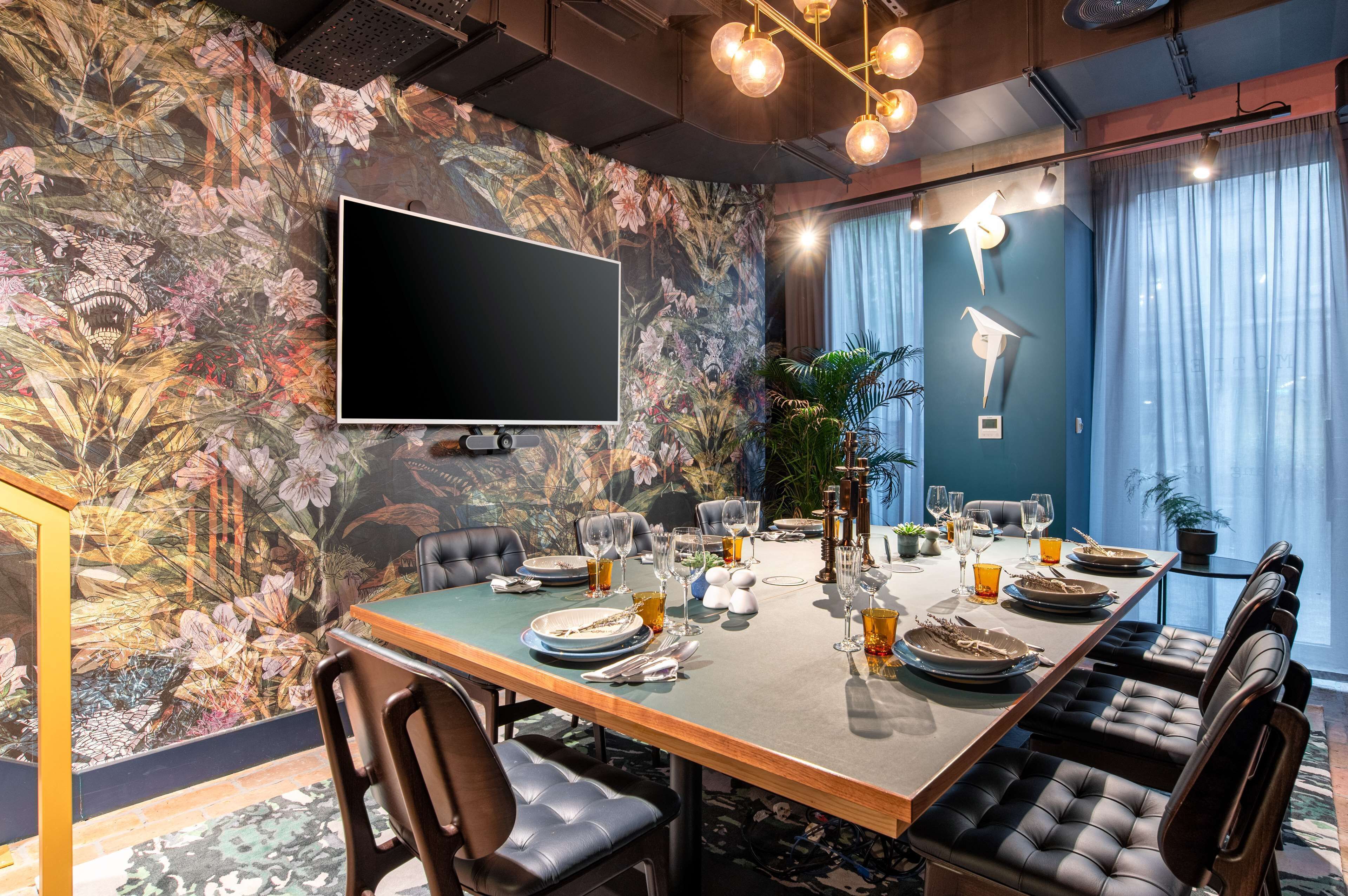 A dining area with a long table set for six, a flat-screen TV mounted on the wall, and a floral mural on the wall.
