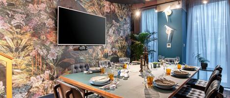 A dining area with a long table set for six, a flat-screen TV mounted on the wall, and a floral mural on the wall.