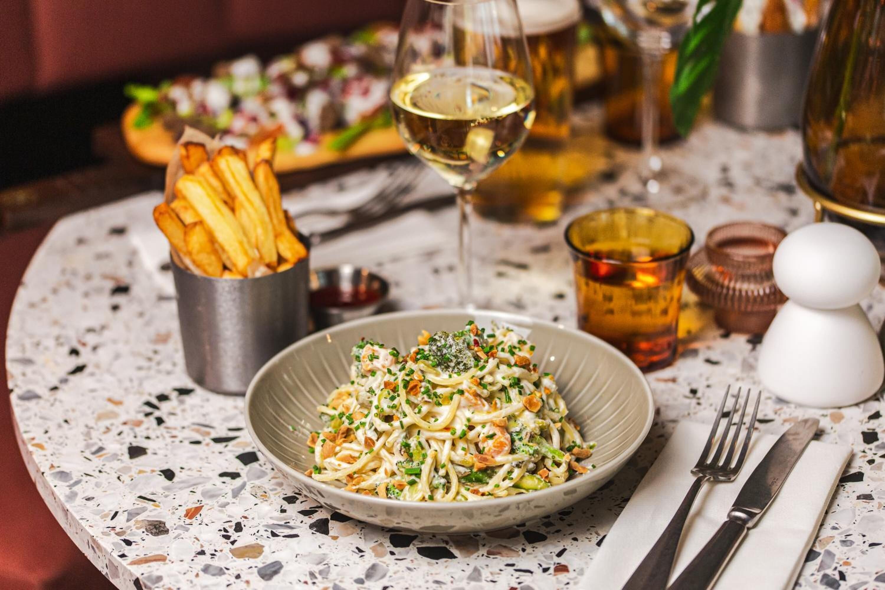 A dining table with a plate of pasta, a glass of white wine, and a small container of fries.