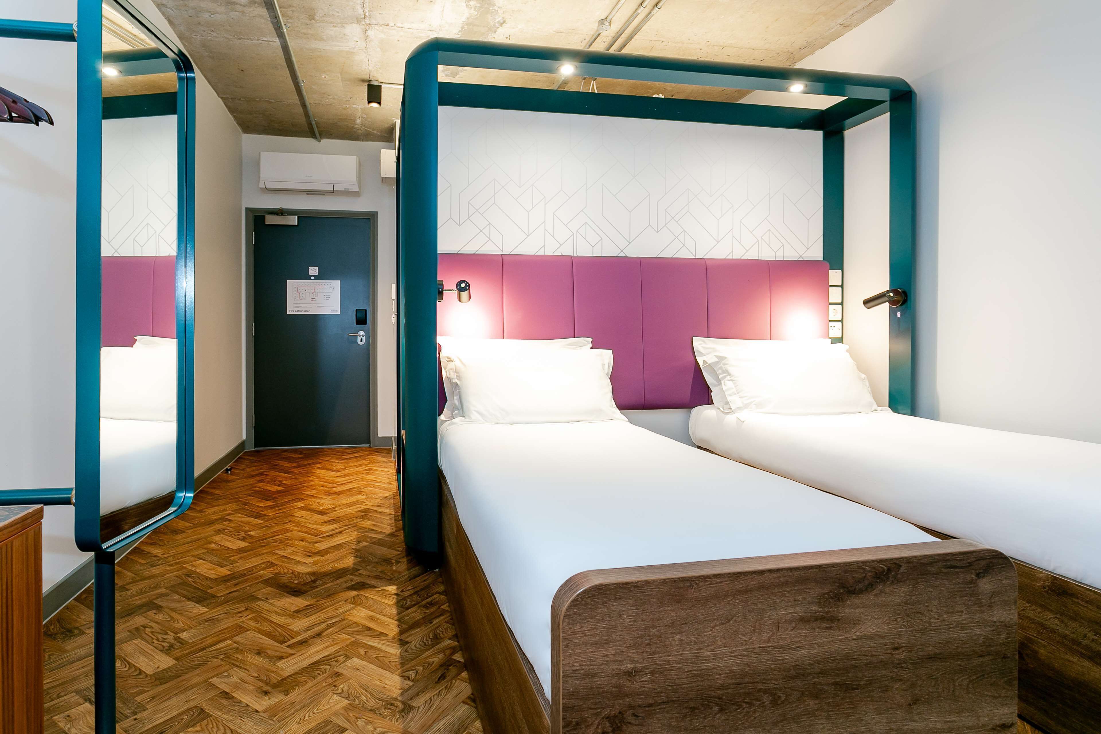A modern hotel room with two single beds, wooden flooring, and a teal-framed canopy bed.