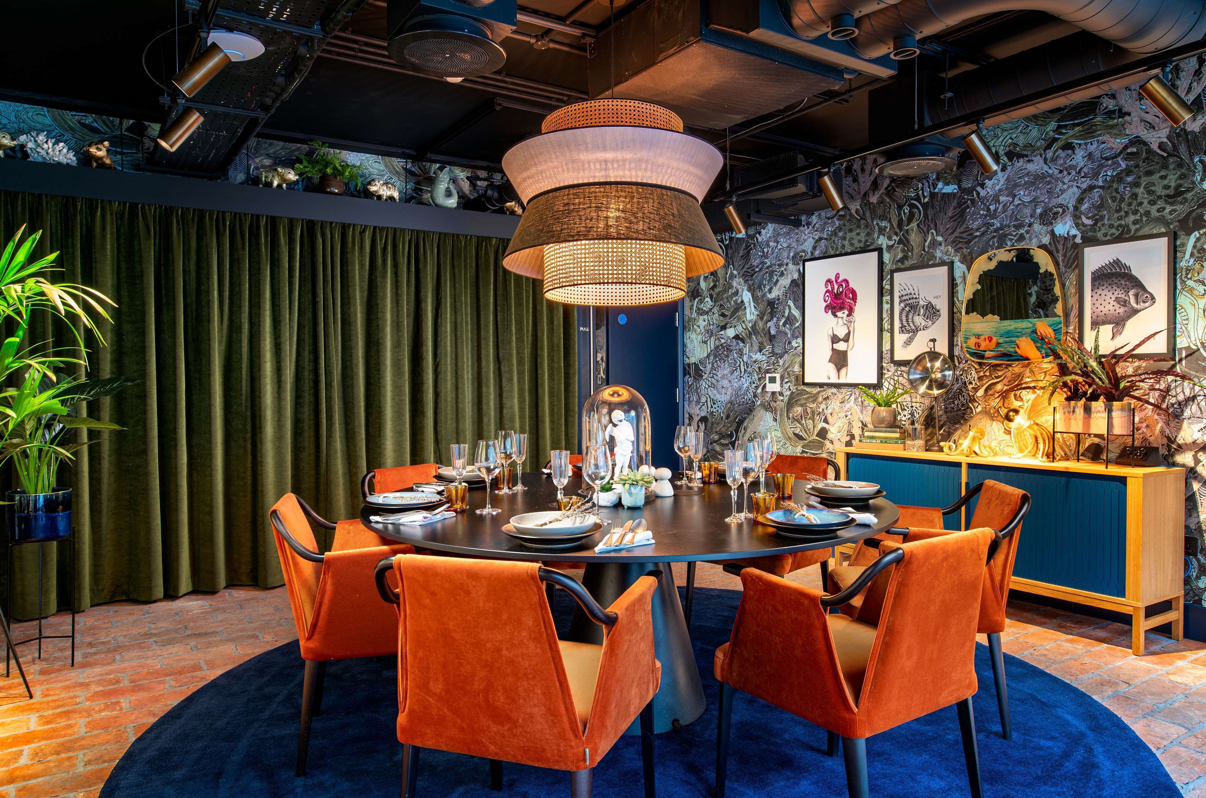 A dining area with a round table set for six, surrounded by orange chairs, with a blue carpet and a green curtain in the background.