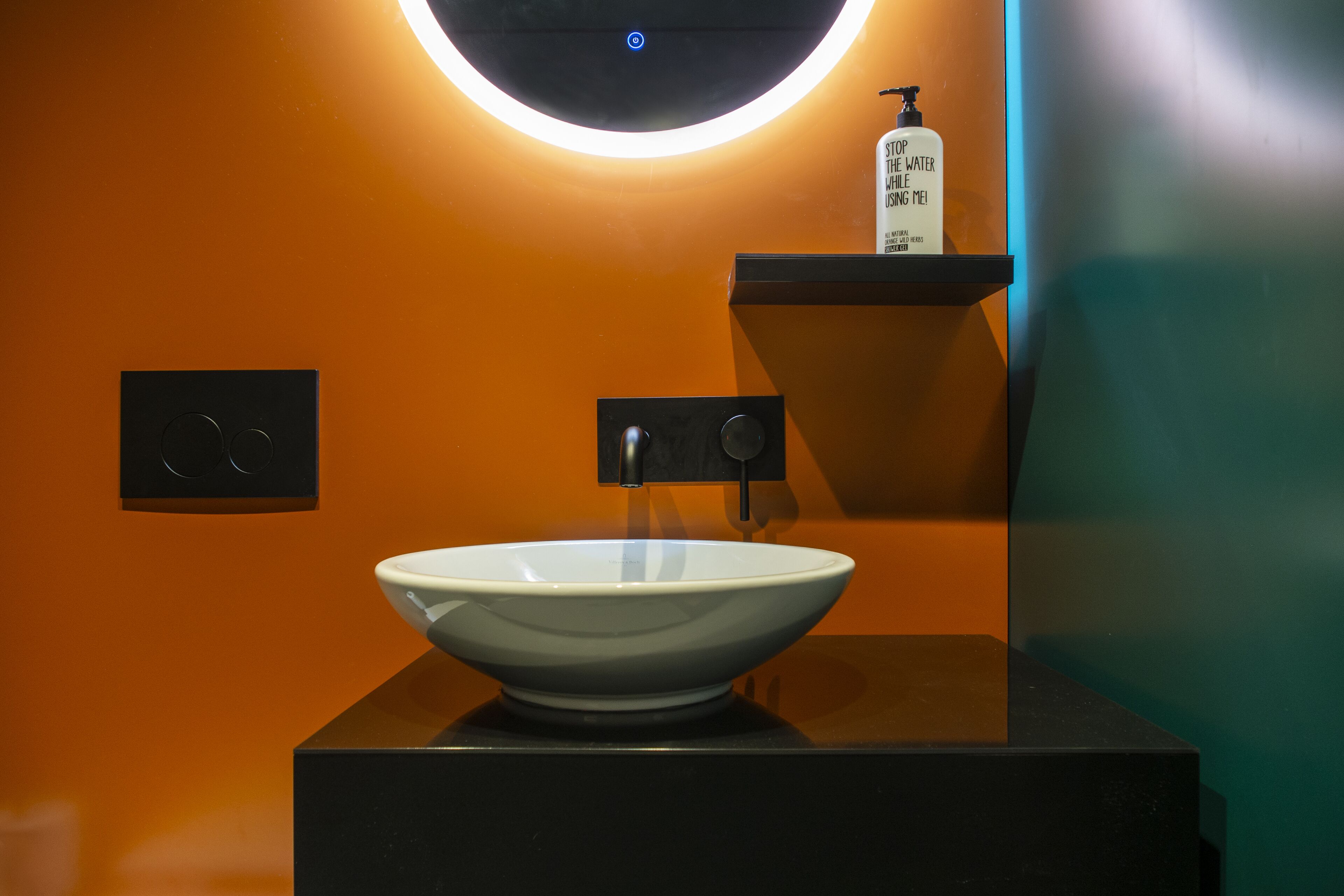 Un baño moderno con un lavamanos de porcelana blanca, un dispensador de jabón y una lámpara fijada a la pared.