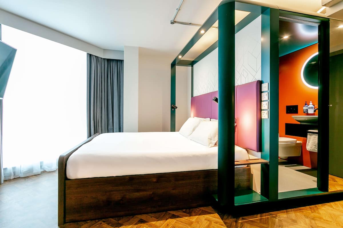 A modern hotel room with a large bed, a bathroom with a circular mirror, and a view of the city.