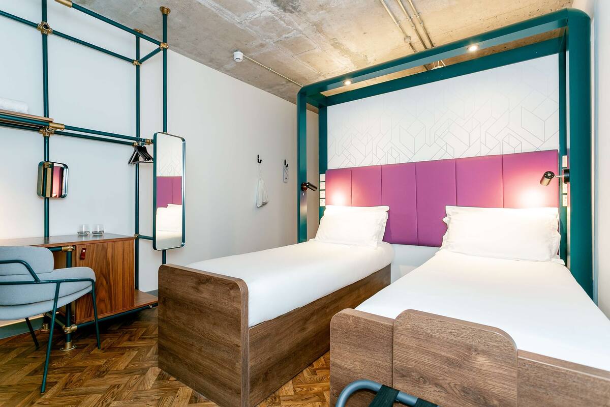 A modern hotel room with two single beds, a wooden desk, a chair, and a herringbone-patterned floor.