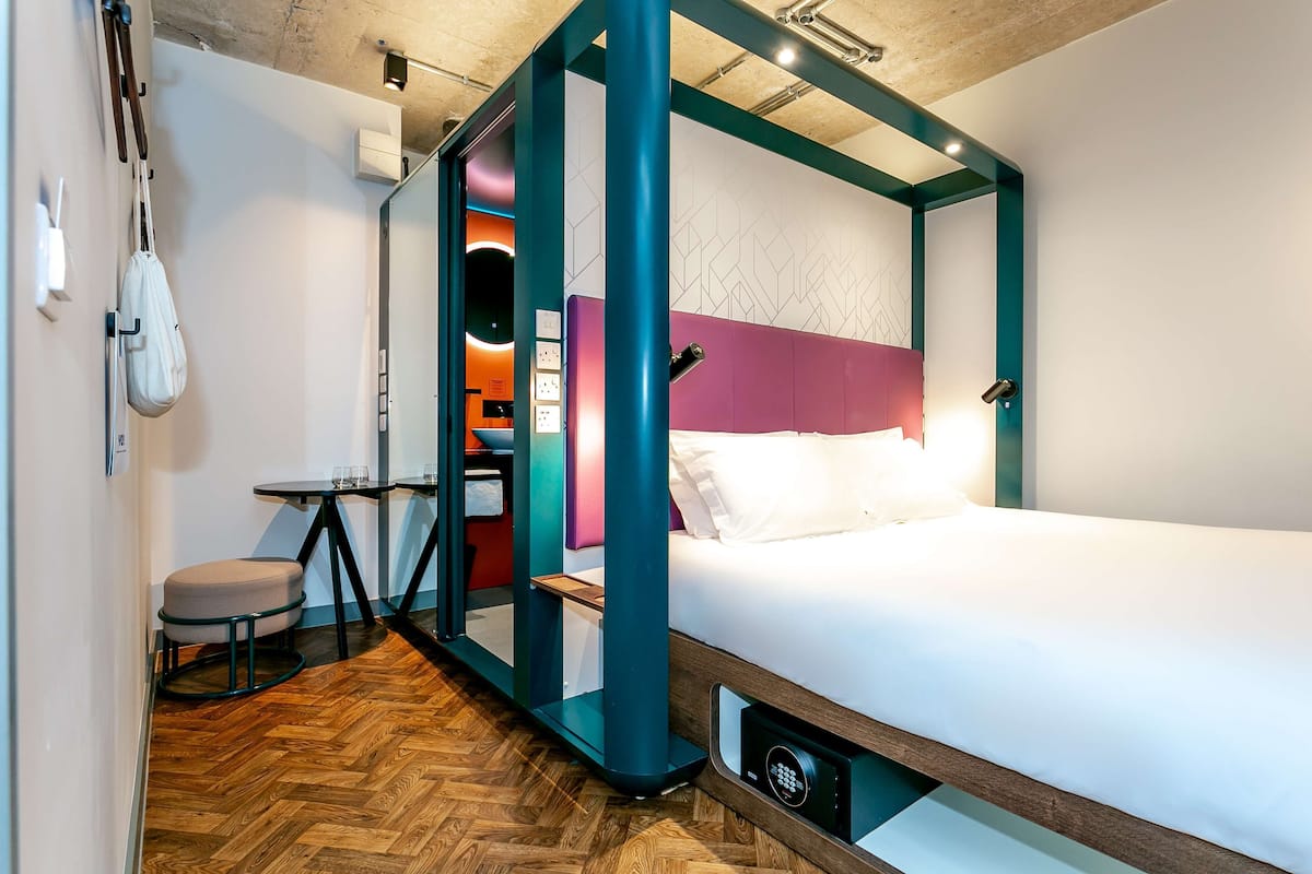 A modern hotel room with a wooden herringbone floor, a bed with a white mattress and teal headboard, a small round table with a chair, and a wall-mounted bag rack.