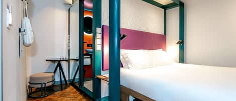 A modern hotel room with a wooden herringbone floor, a bed with a white mattress and teal headboard, a small round table with a chair, and a wall-mounted bag rack.