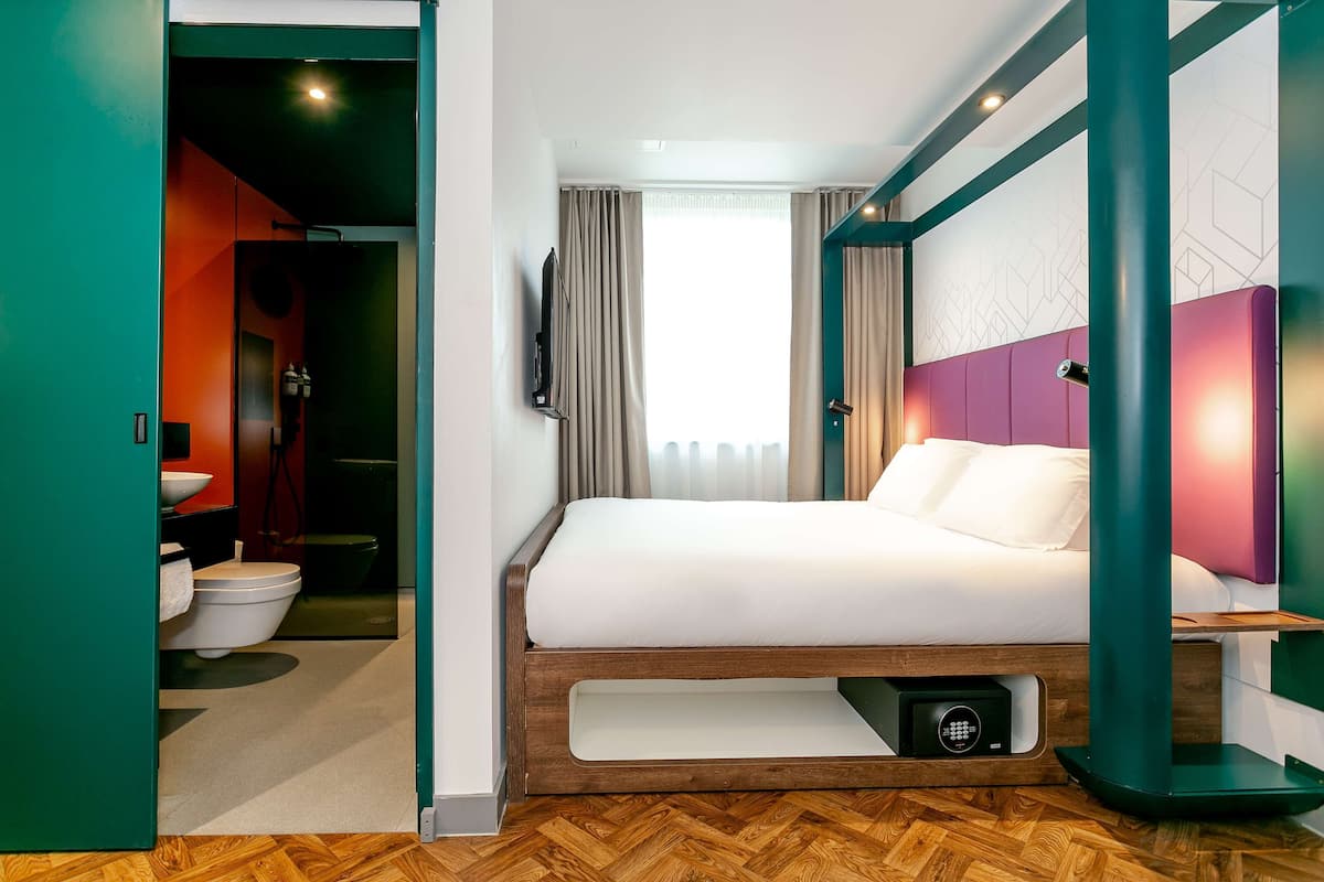 A modern hotel room with a wooden floor, a bed with white bedding, a TV mounted on the wall, and a bathroom visible through an open door.