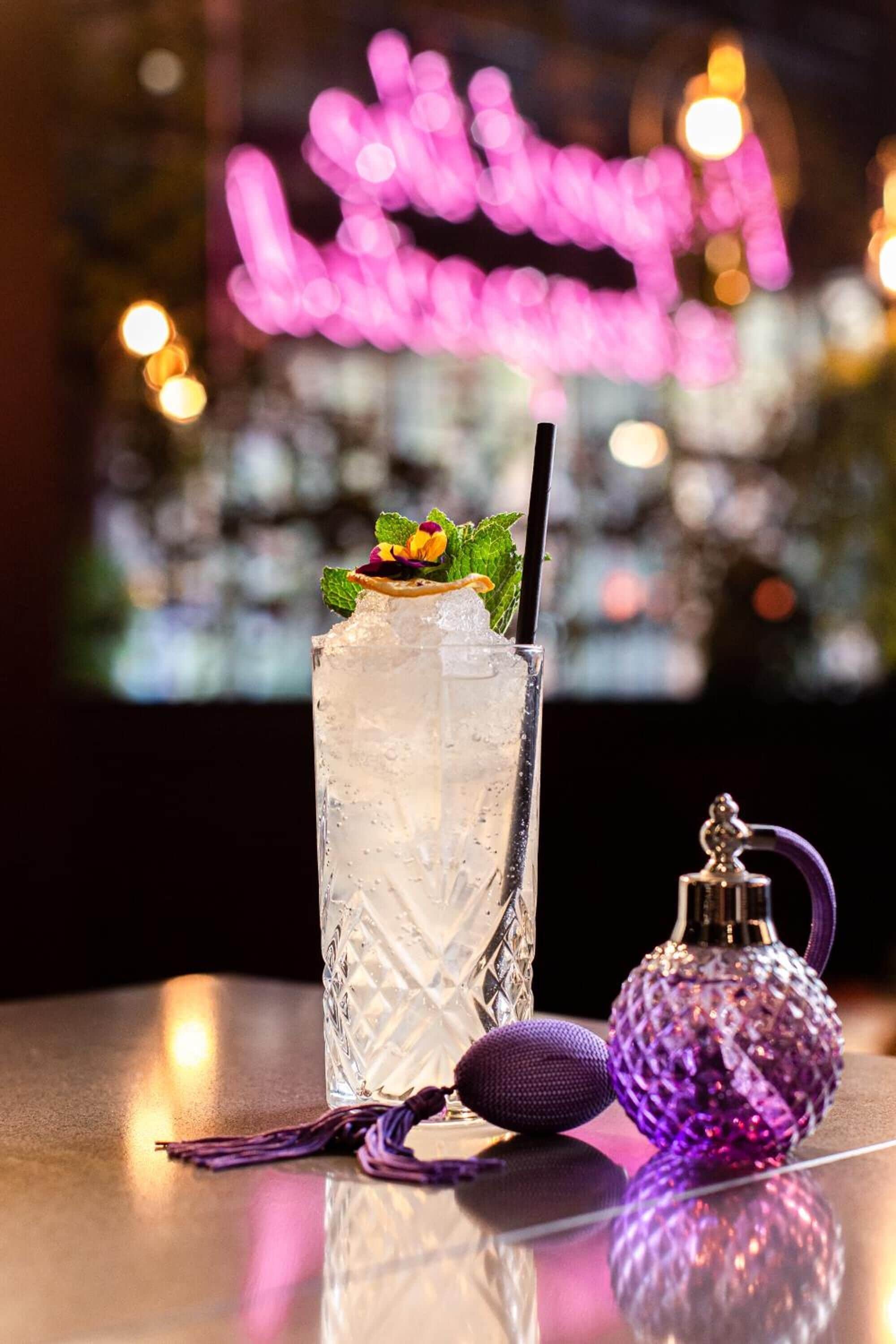 A tall glass with a drink, a purple decorative item, and a small bottle on a reflective surface.