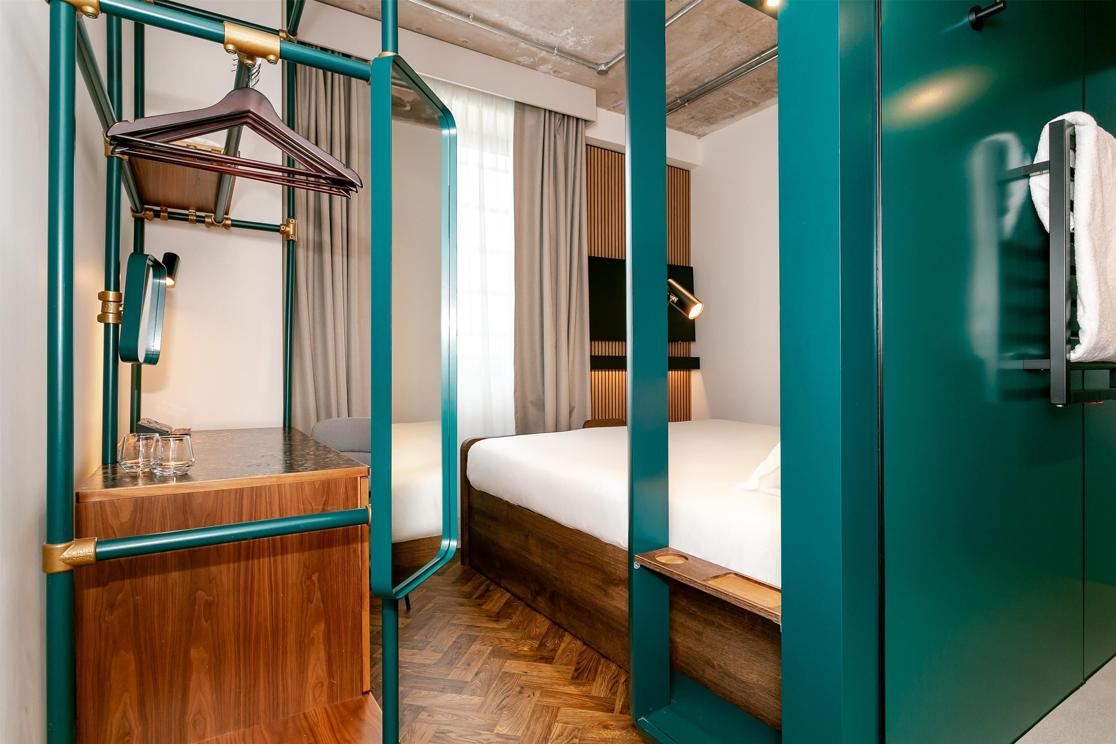 A compact hotel room with a wooden dresser, a bed with white linens, teal-colored walls, and a glass shower enclosure.