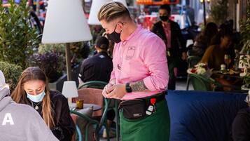 A waiter in a pink shirt and green apron standing at an outdoor cafe with tables and chairs.