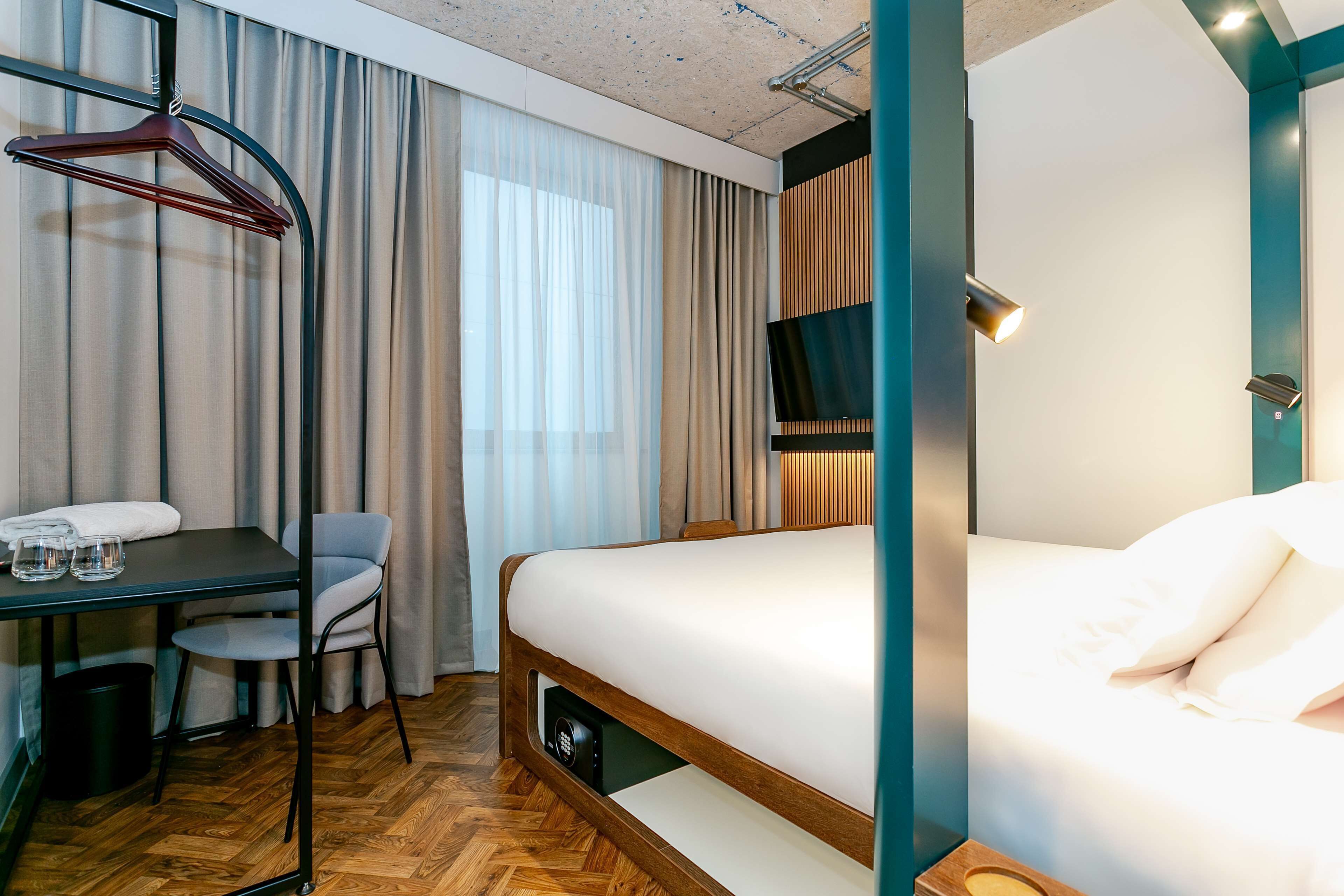 Una camera d'albergo moderna con pavimento in legno, un letto con lenzuola bianche, una scrivania con sedia, una TV montata a parete e un appendiabiti con grucce.