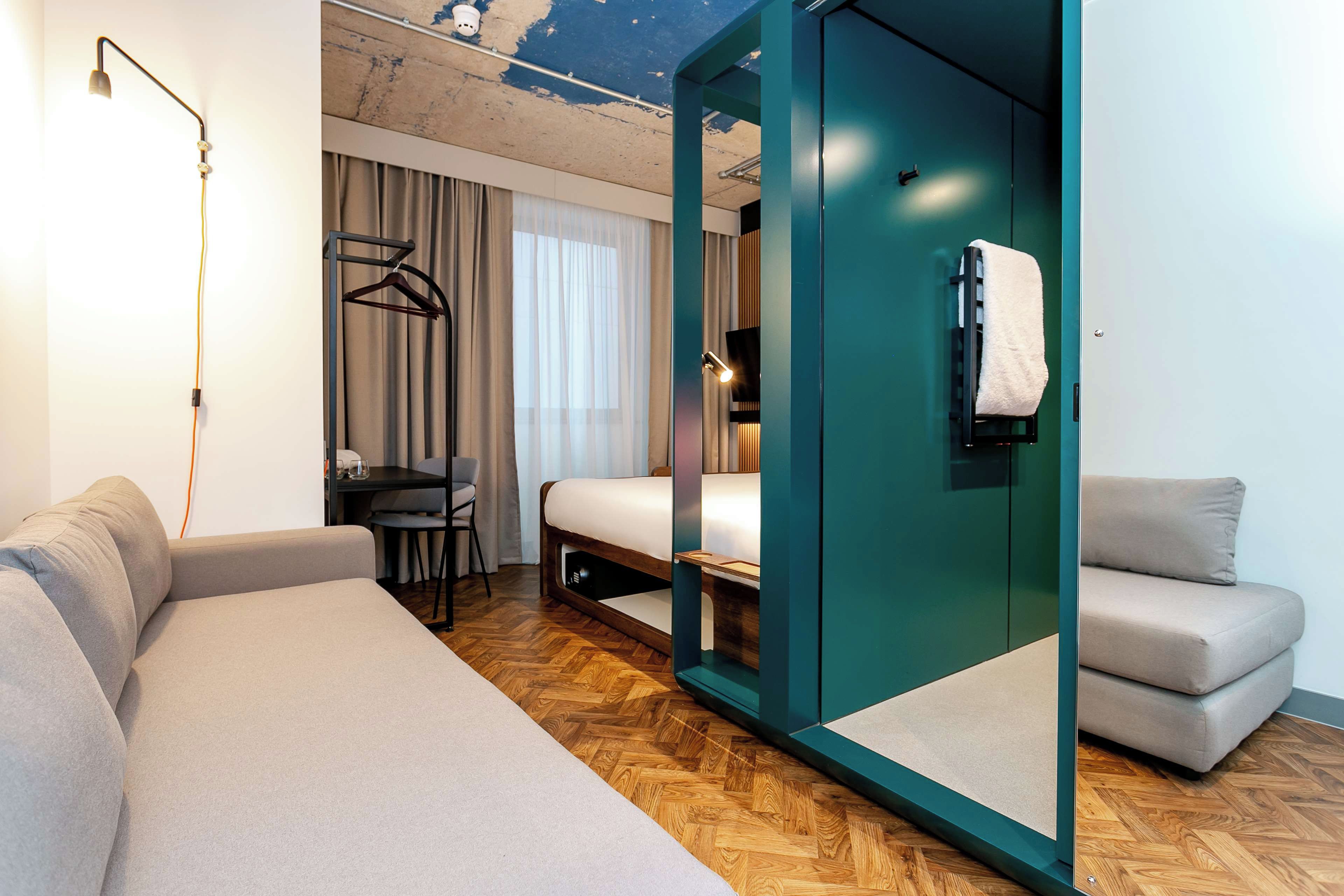 Ett modernt hotellrum med en säng, ett skrivbord, en stol, en soffa och en spegel.