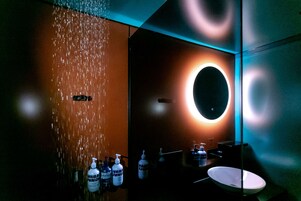 Un baño moderno con ducha de lluvia, un espejo redondo y varias botellas de gel hidroalcohólico.