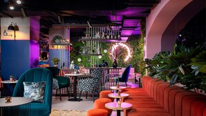 Un'area lounge moderna con sedute color verde acqua e arancione, una parete verde con piante e un angolo bar con insegna al neon.