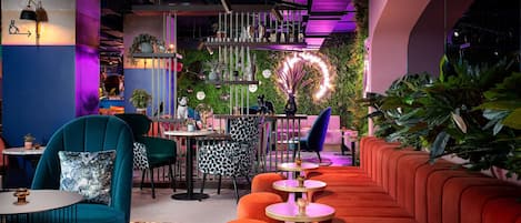 A modern lounge area with teal and orange seating, a green wall with plants, and a bar area with a neon sign.