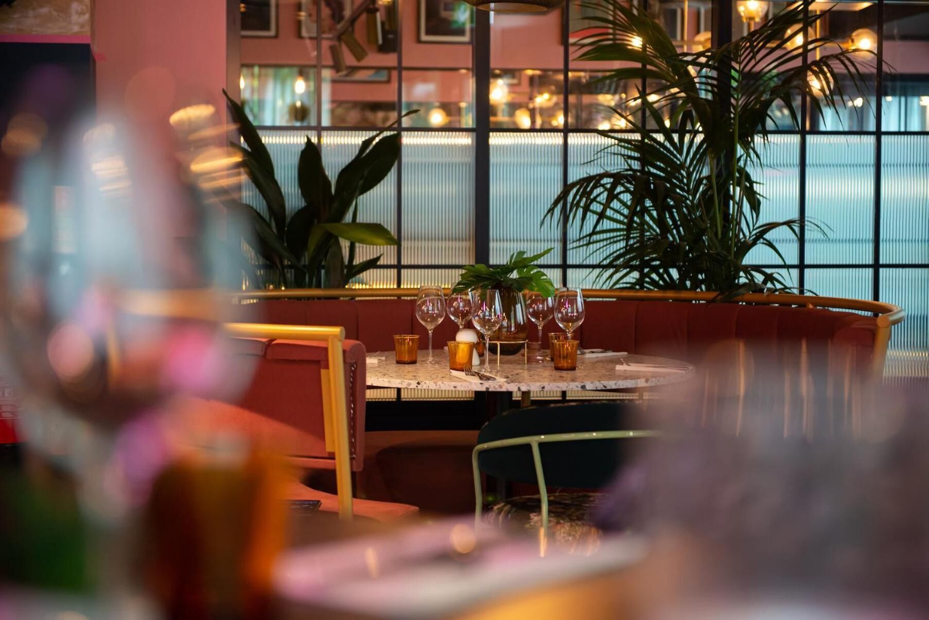Ein mit Weingläsern und einer Pflanze auf einem Tisch gedeckter Restauranttisch, mit Blick auf ein Gebäude durch große Fenster.